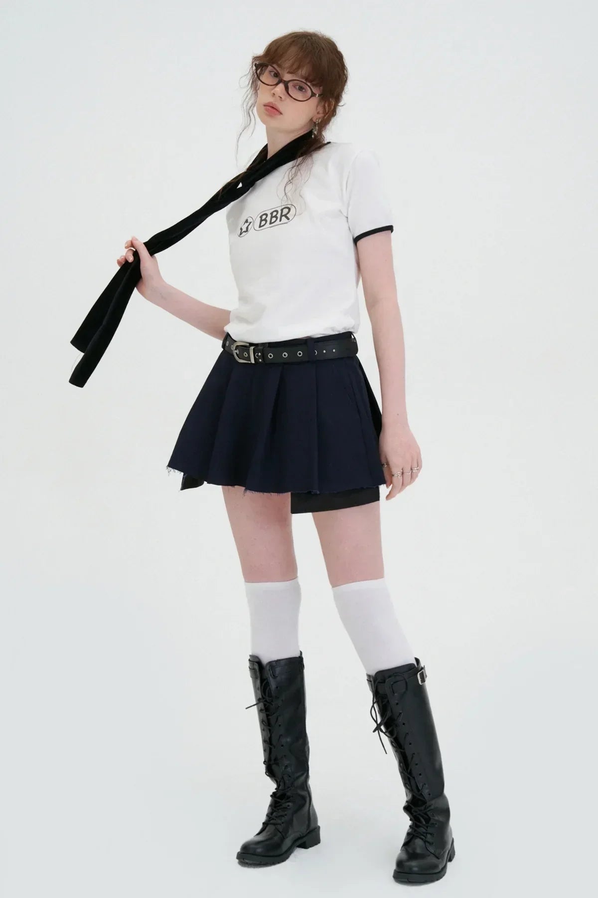 Woman wearing navy wide tuck mini skirt with black belt, white graphic tee, knee-high black lace-up boots, and white knee socks