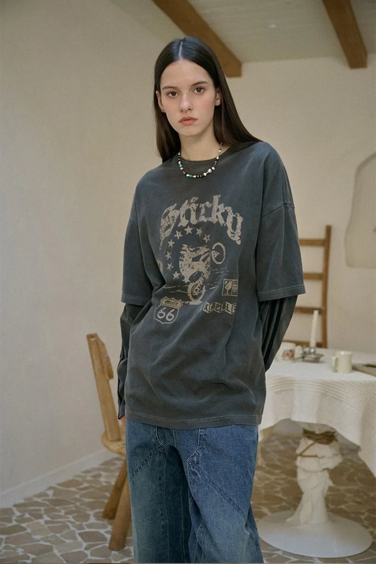 Young woman wearing a dark layered long-sleeve shirt with graphic print and blue jeans indoors