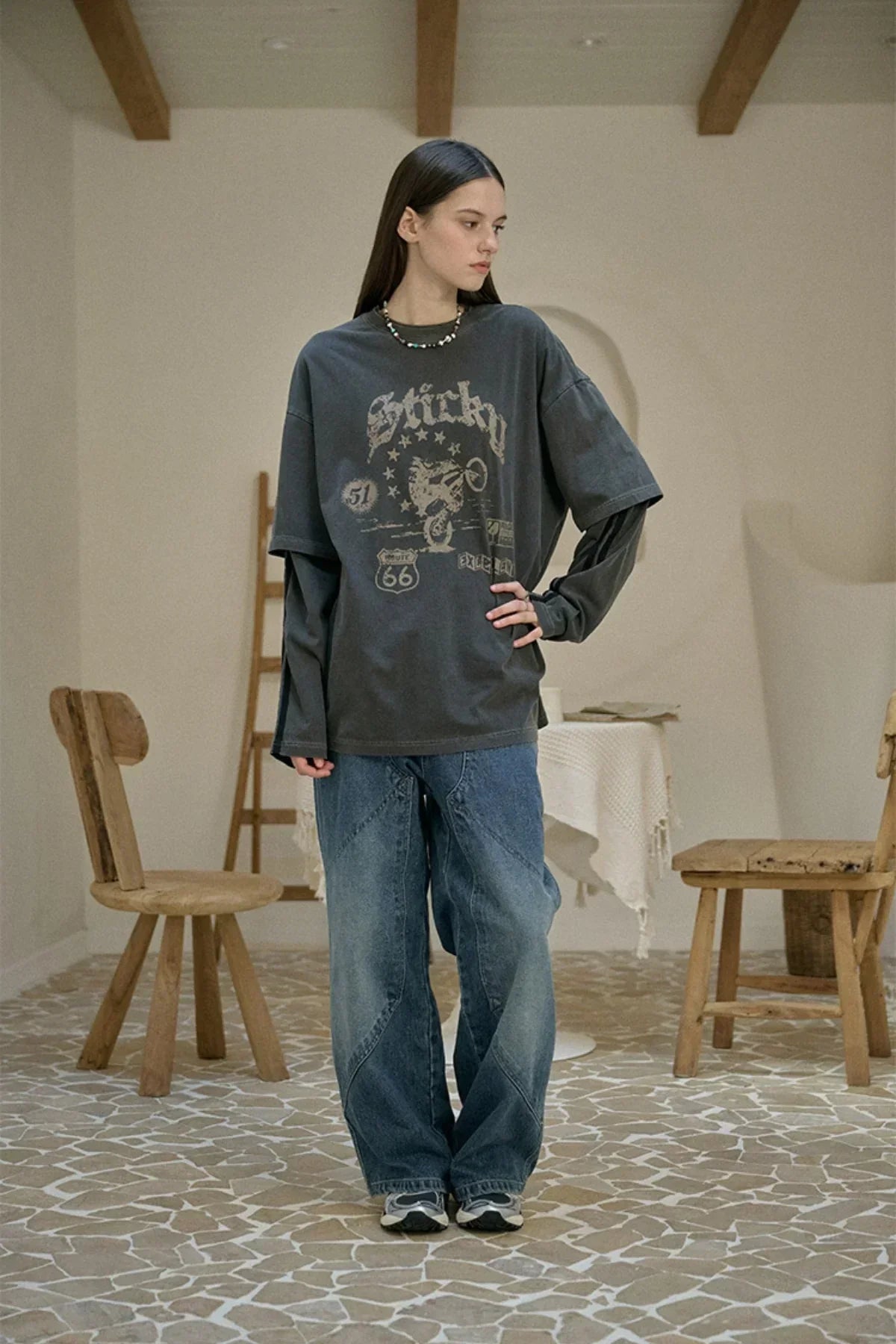 Model wearing layered long sleeve gray graphic t-shirt and loose blue jeans in rustic room with wooden chairs