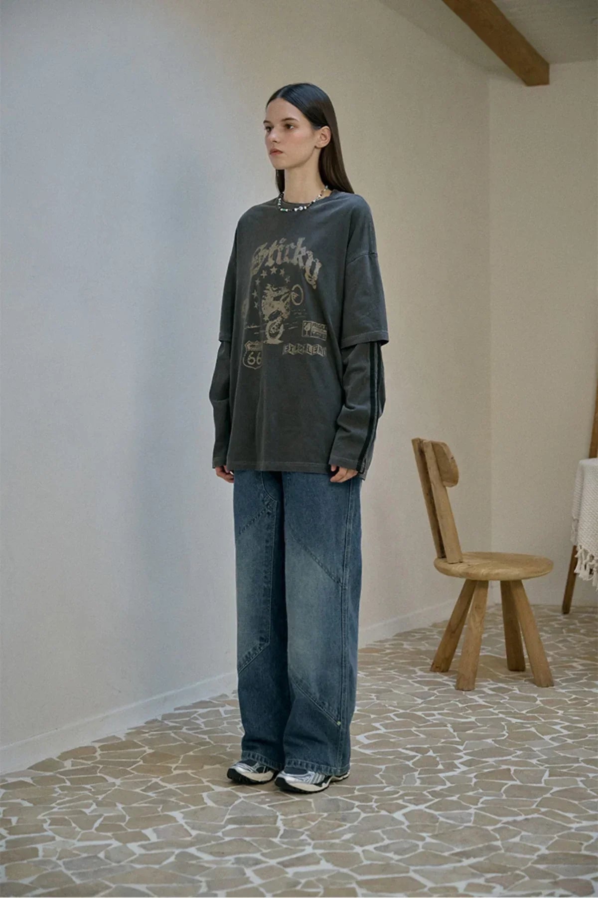 Young woman wearing layered smoky black long sleeve graphic t-shirt and wide denim jeans standing indoors