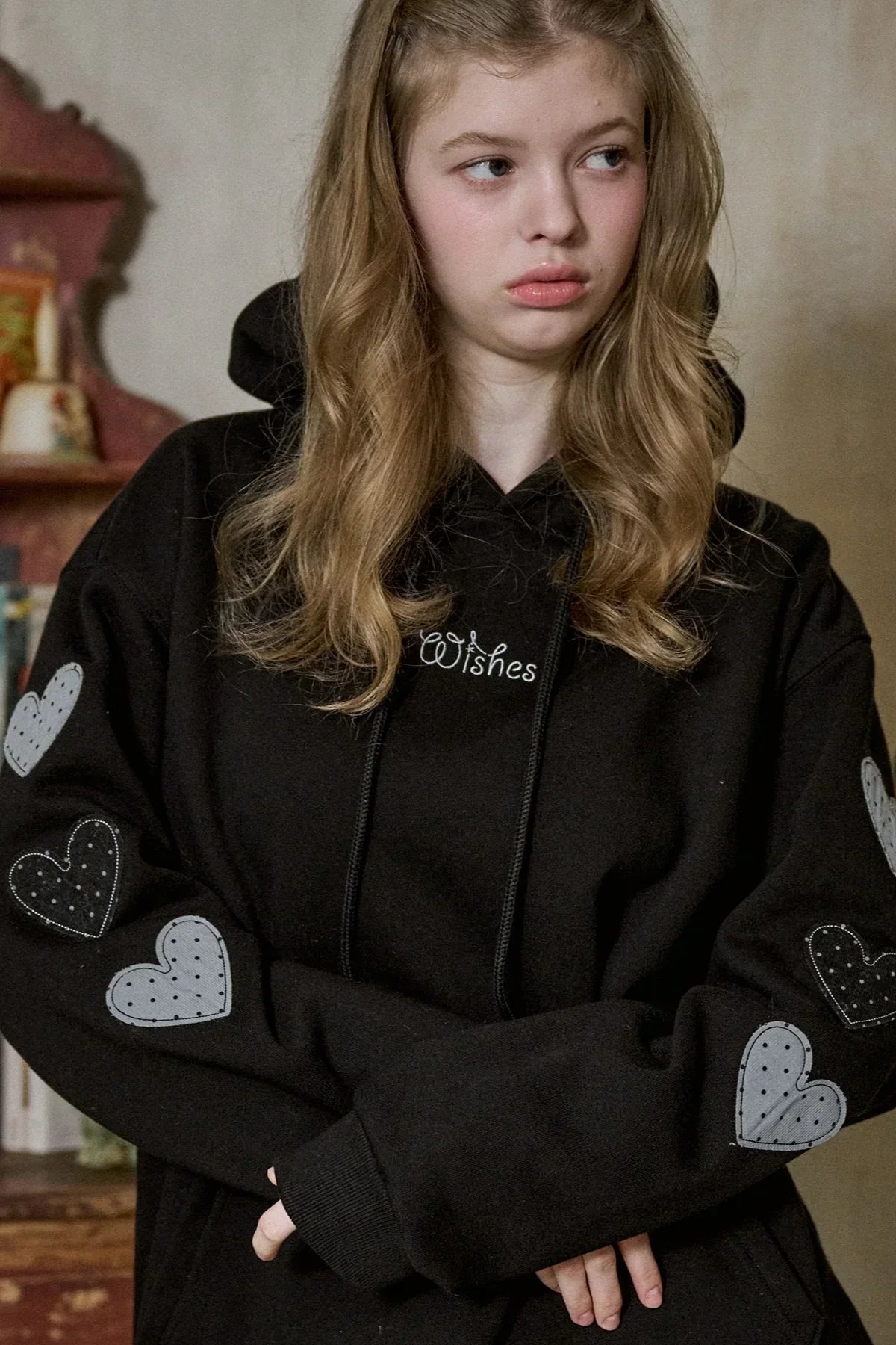Young woman wearing black hoodie with stitched heart patches on sleeves and "Wishes" text on chest