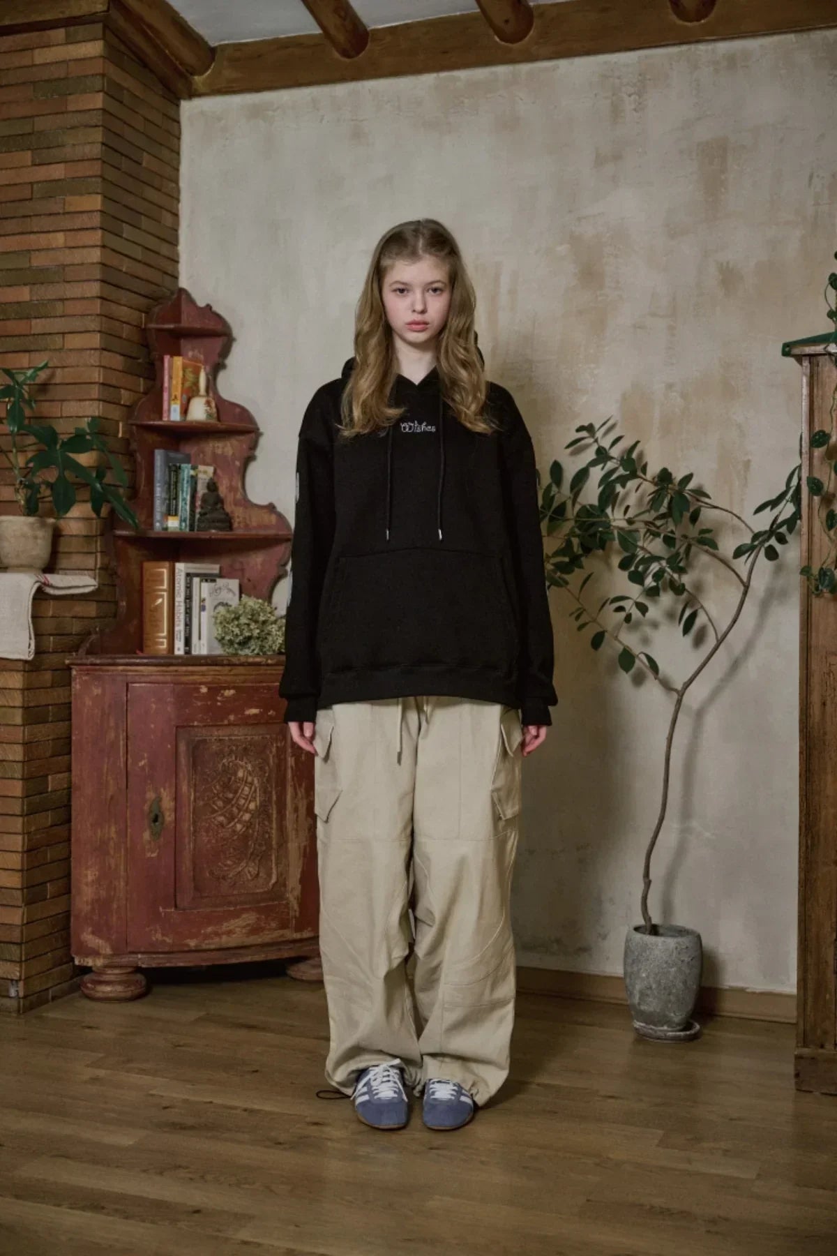 Young woman in black hoodie and beige cargo pants standing indoors with rustic wooden furniture and potted plant