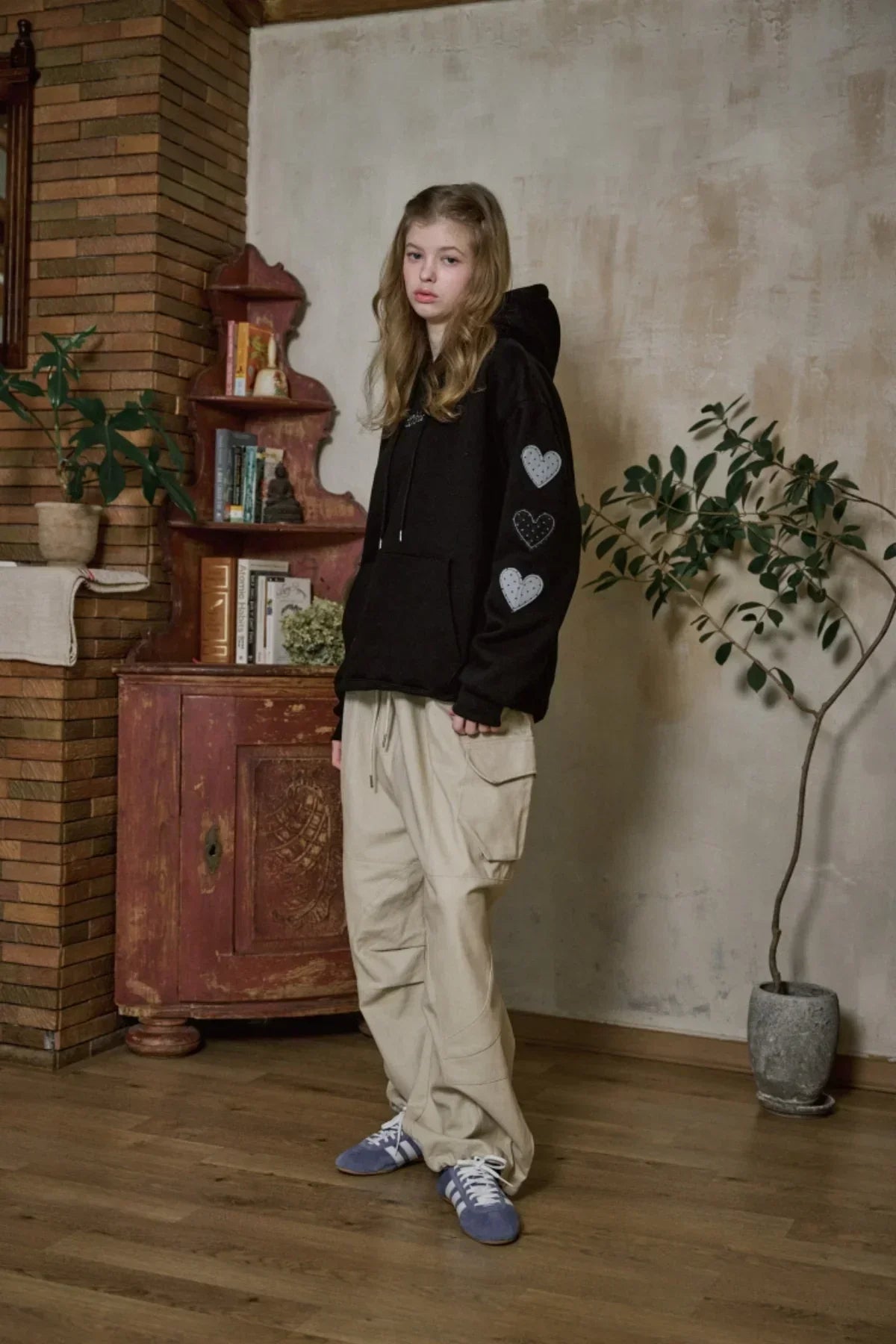 Young woman wearing black hoodie with heart patches and beige cargo pants in a rustic indoor setting