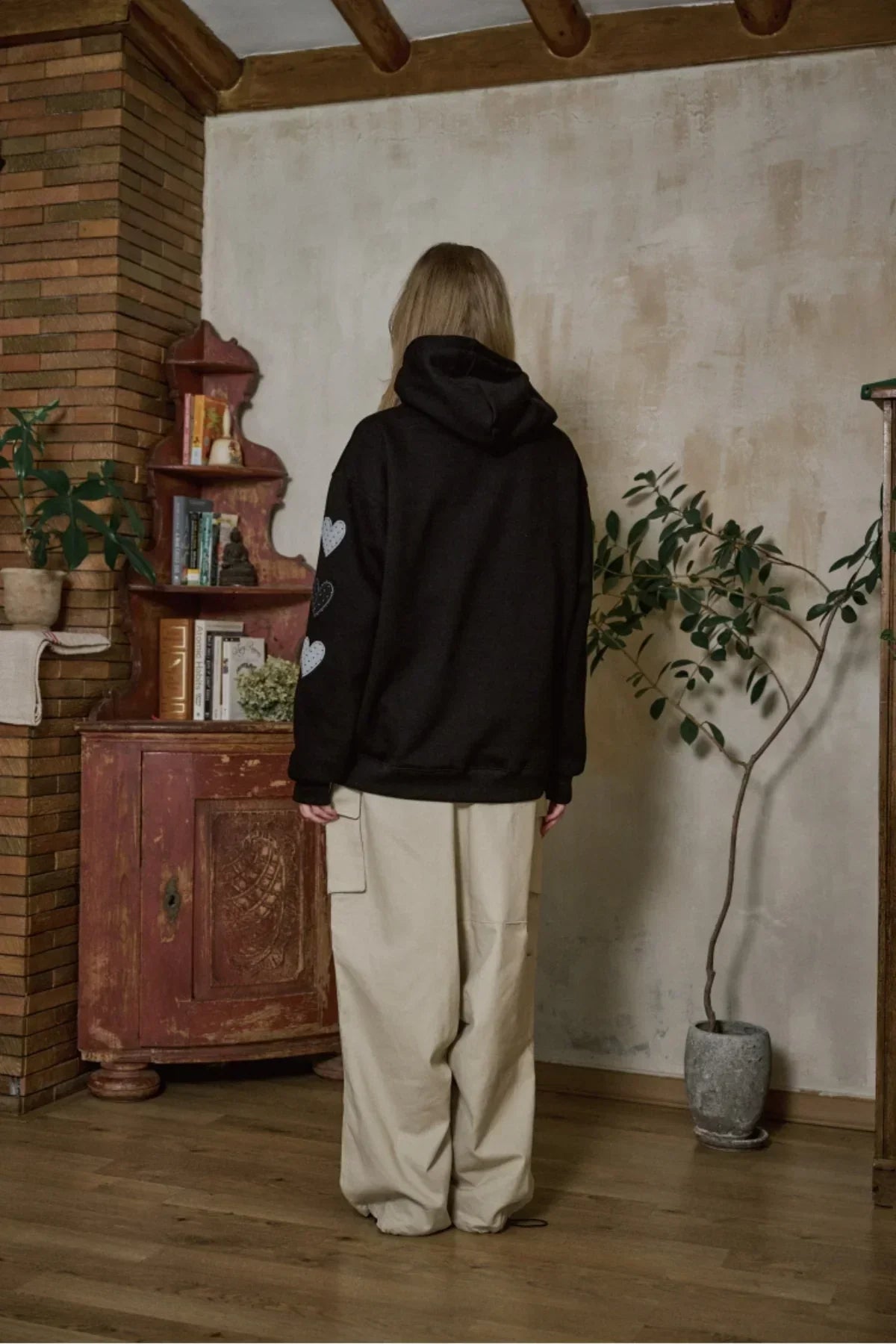 Model wearing black corduroy hoodie with heart patches, paired with beige wide-leg pants in cozy room