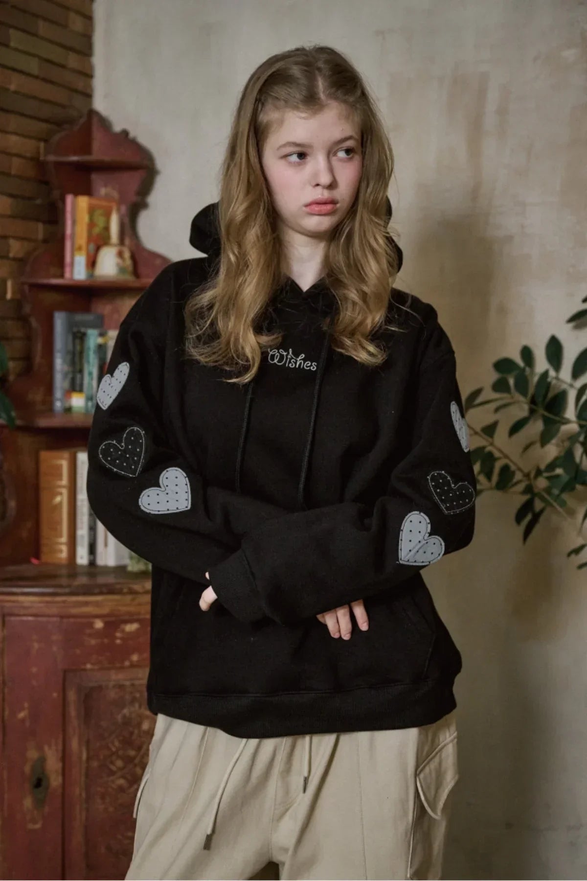 Young woman wearing black hoodie with grey heart patches and Wishes embroidery in cozy indoor setting