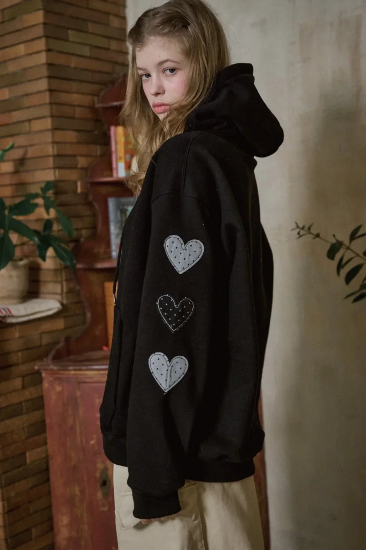 Young woman wearing black hoodie with polka dot heart patches on sleeve in cozy indoor setting