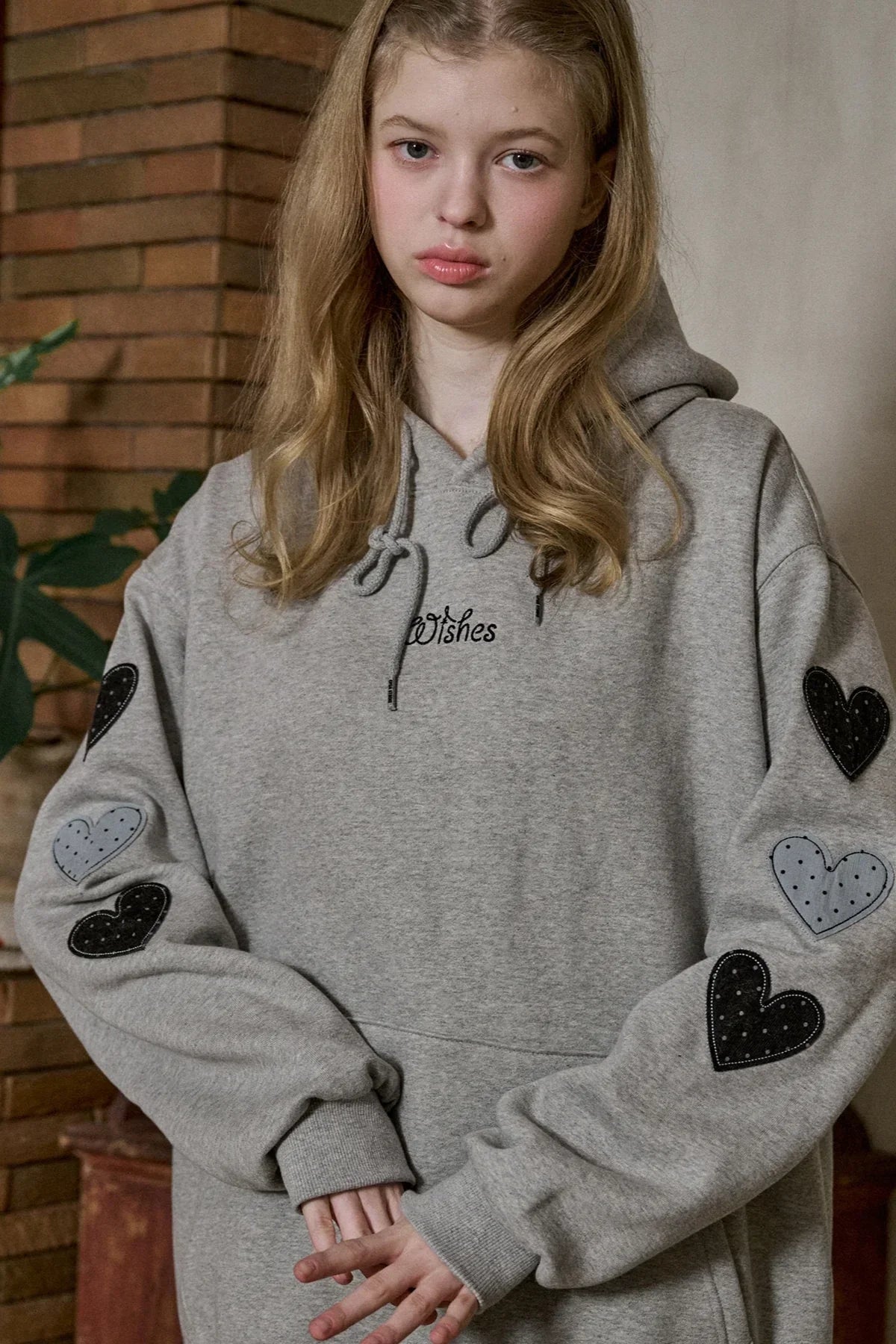 Young woman wearing gray hoodie with heart patches on sleeves and "Wishes" text on chest