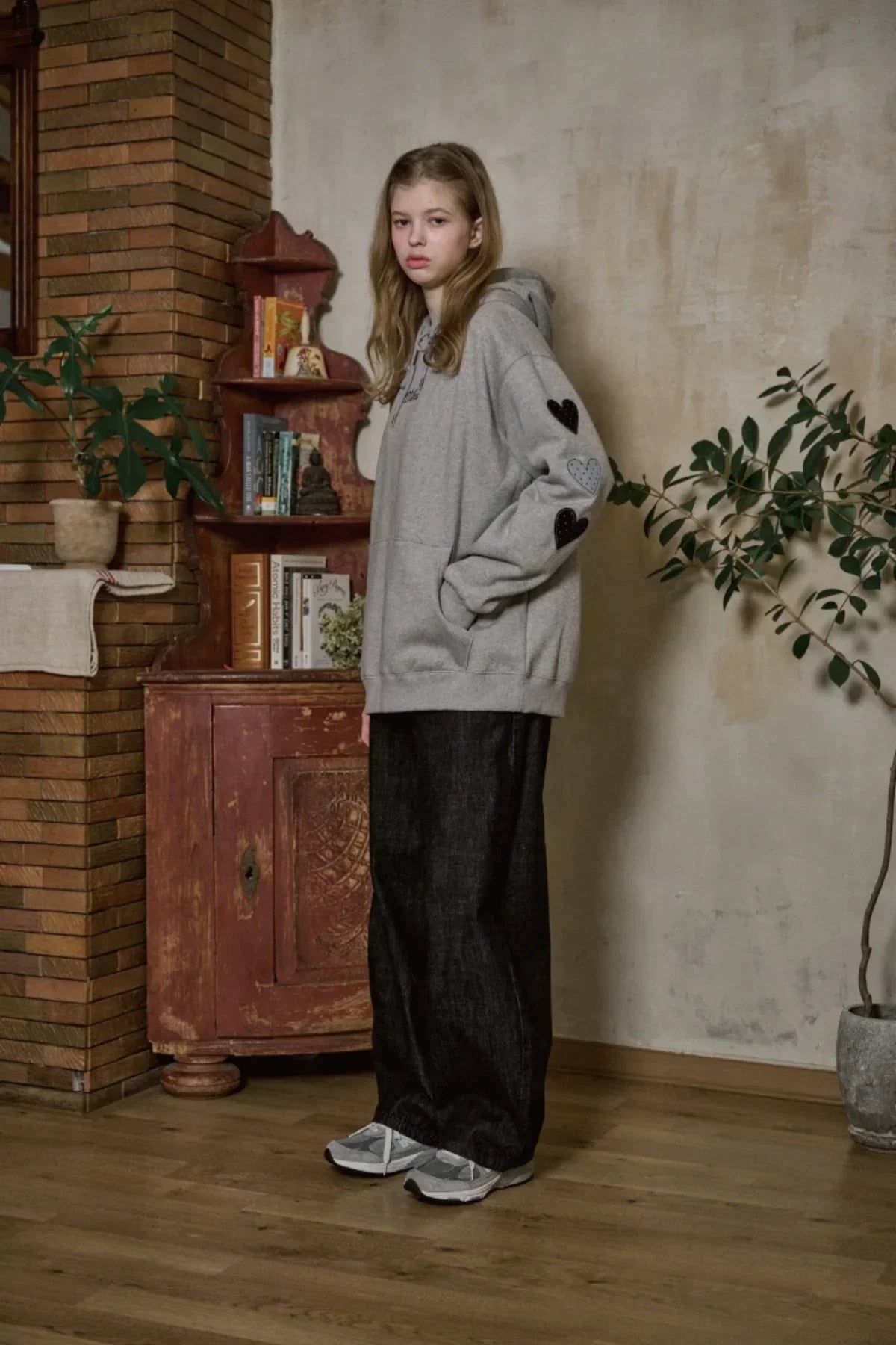 Model wearing gray hoodie with heart patches, wide black pants, and sneakers in cozy room