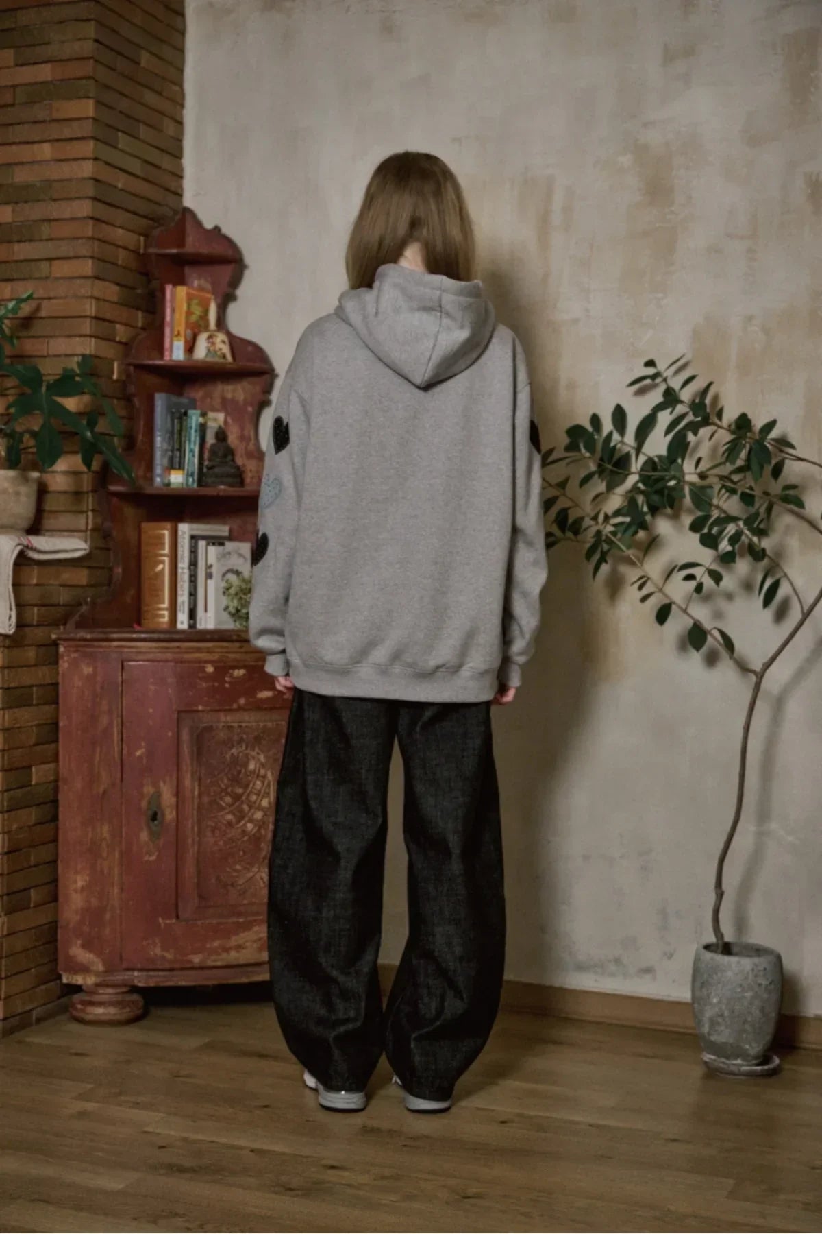 Person wearing gray hoodie with heart patches and loose black pants standing indoors by vintage cabinet and plant