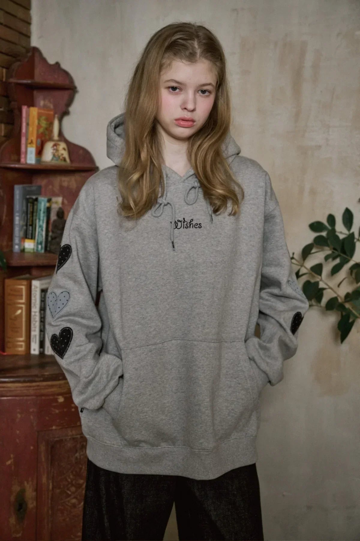 Young woman wearing gray heart-patch hoodie with text, standing indoors near vintage bookshelf and green plant