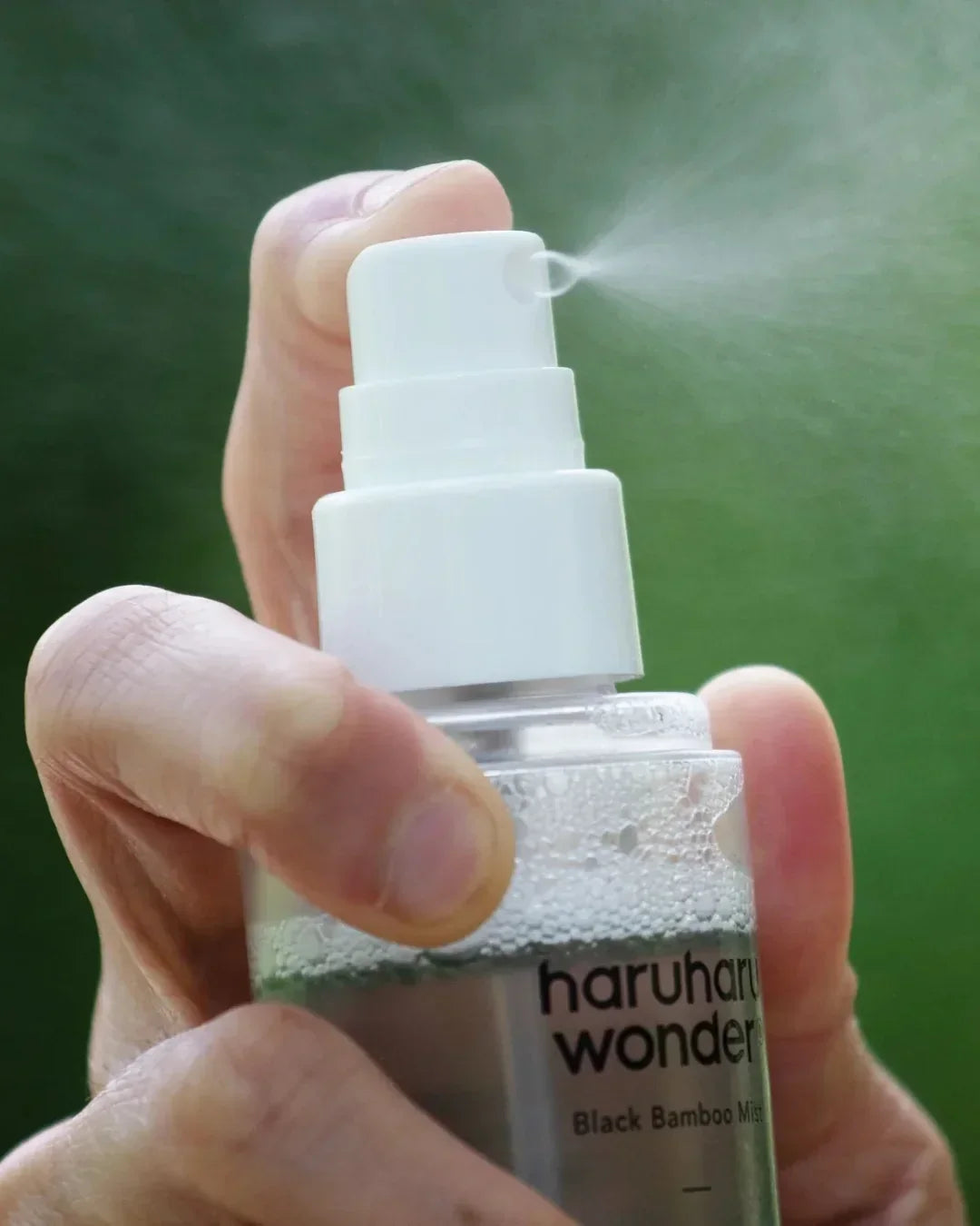 Hand spraying Haruharu Wonder Black Bamboo Mist with white spray nozzle against green background