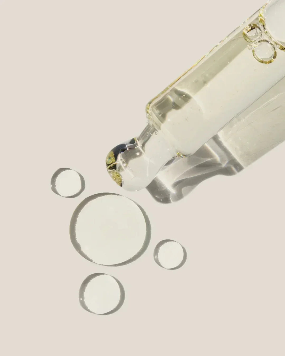 Close-up of clear facial oil dropper releasing transparent oil droplets on beige background