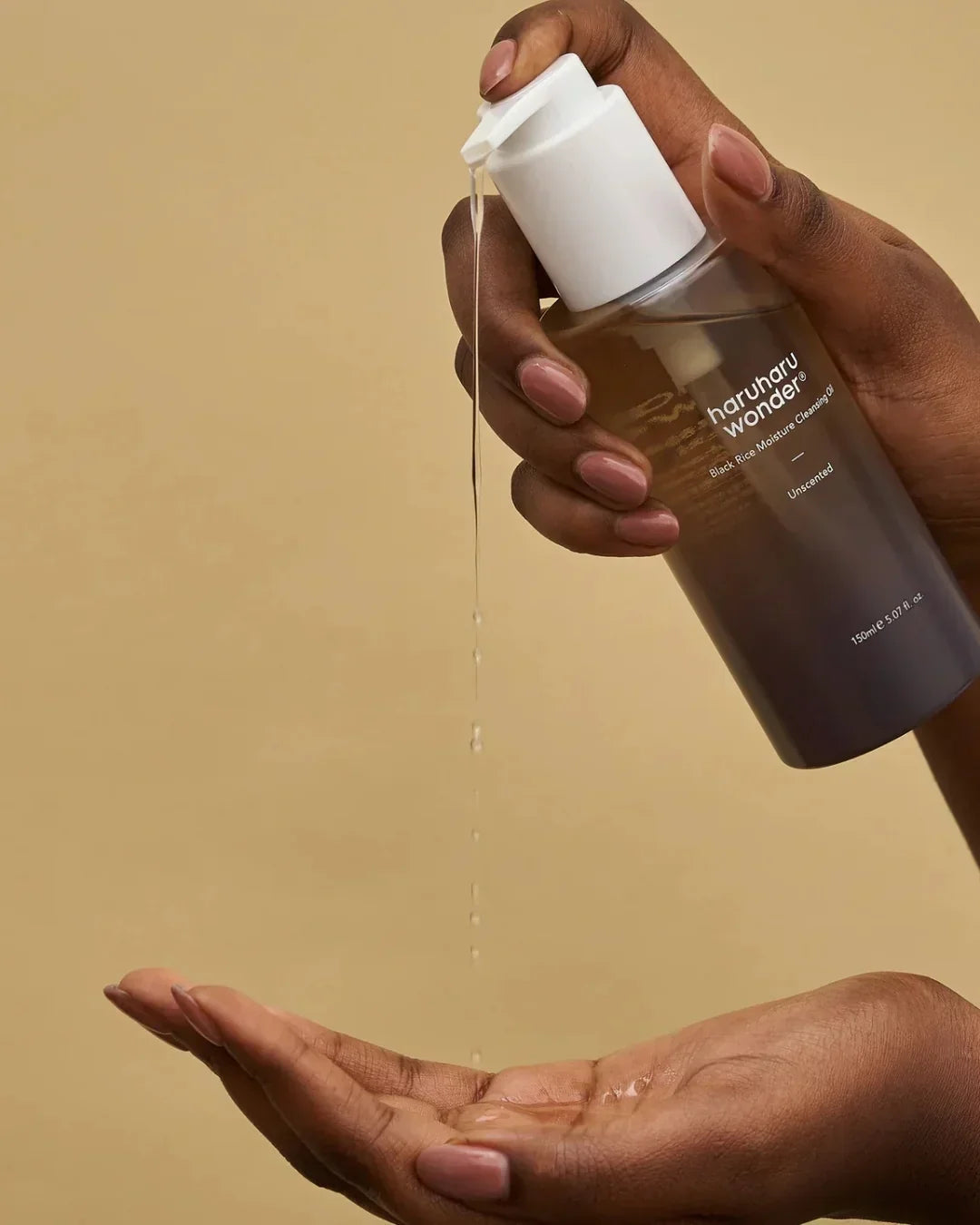 Hands dispensing Haruharu Wonder Black Rice Moisture Cleansing Oil with liquid drops against beige background