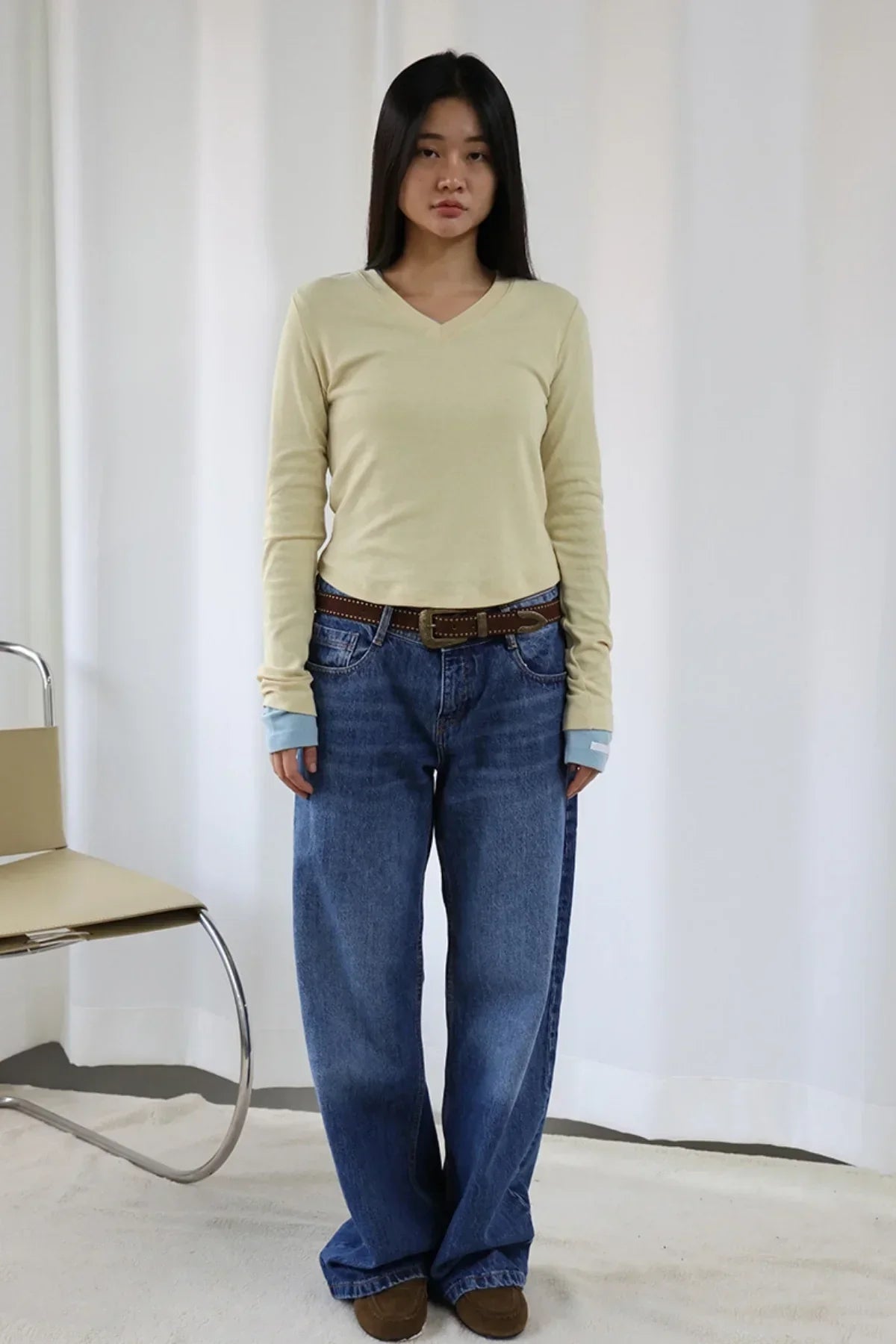 Woman wearing a light yellow V-neck long sleeve tee, layered with blue cuffs, blue wide-leg jeans, brown belt, and brown shoes