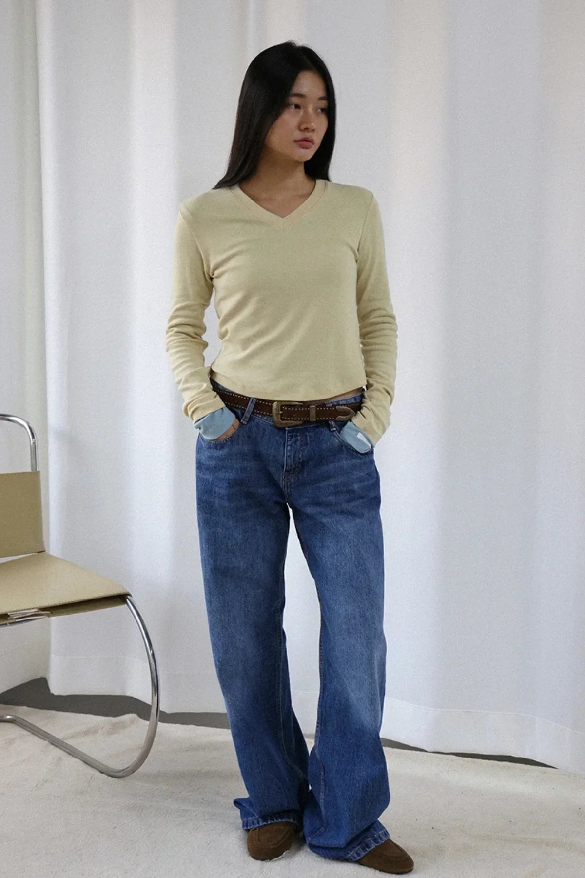 Woman wearing beige V-neck long sleeve wool tee, blue jeans, brown belt, standing near chair in white room