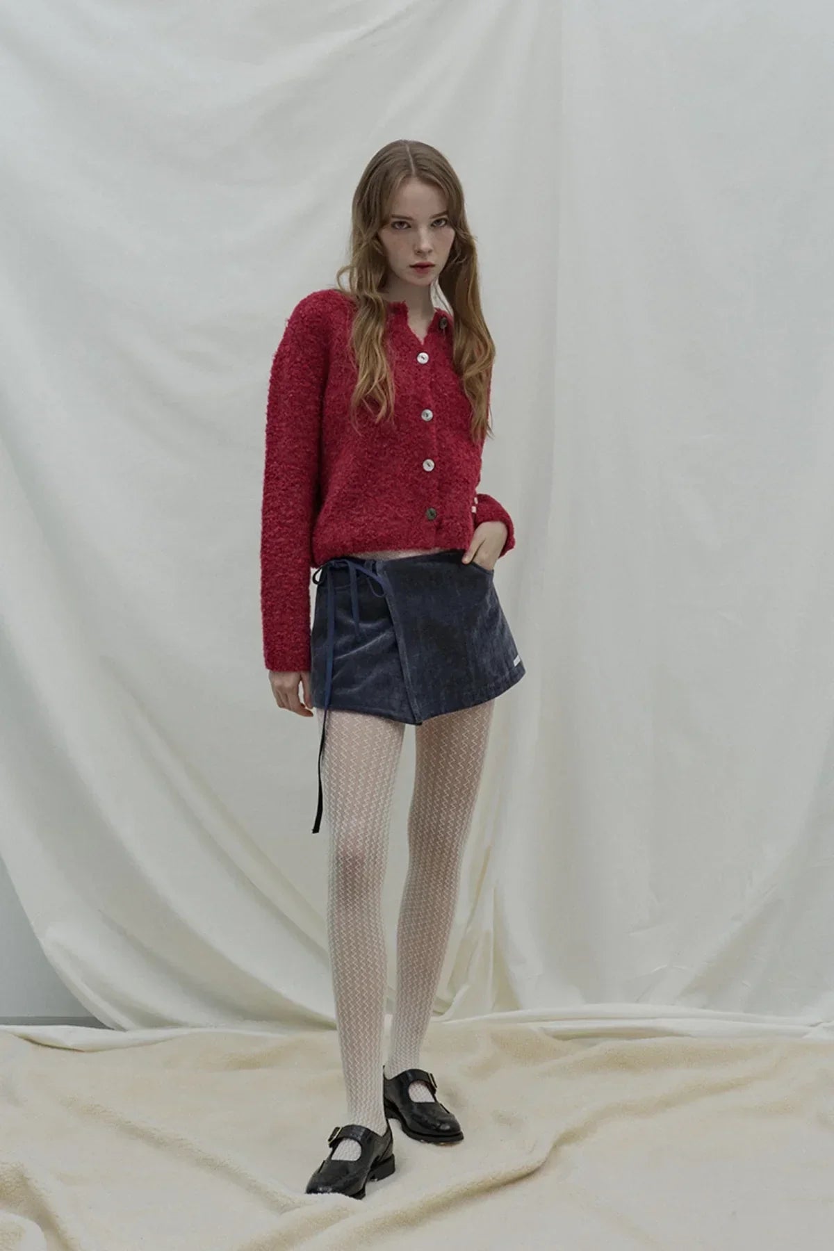 Model wearing a red fuzzy cardigan, navy blue wrap skirt, patterned white tights, and black Mary Jane shoes against a light fabric backdrop