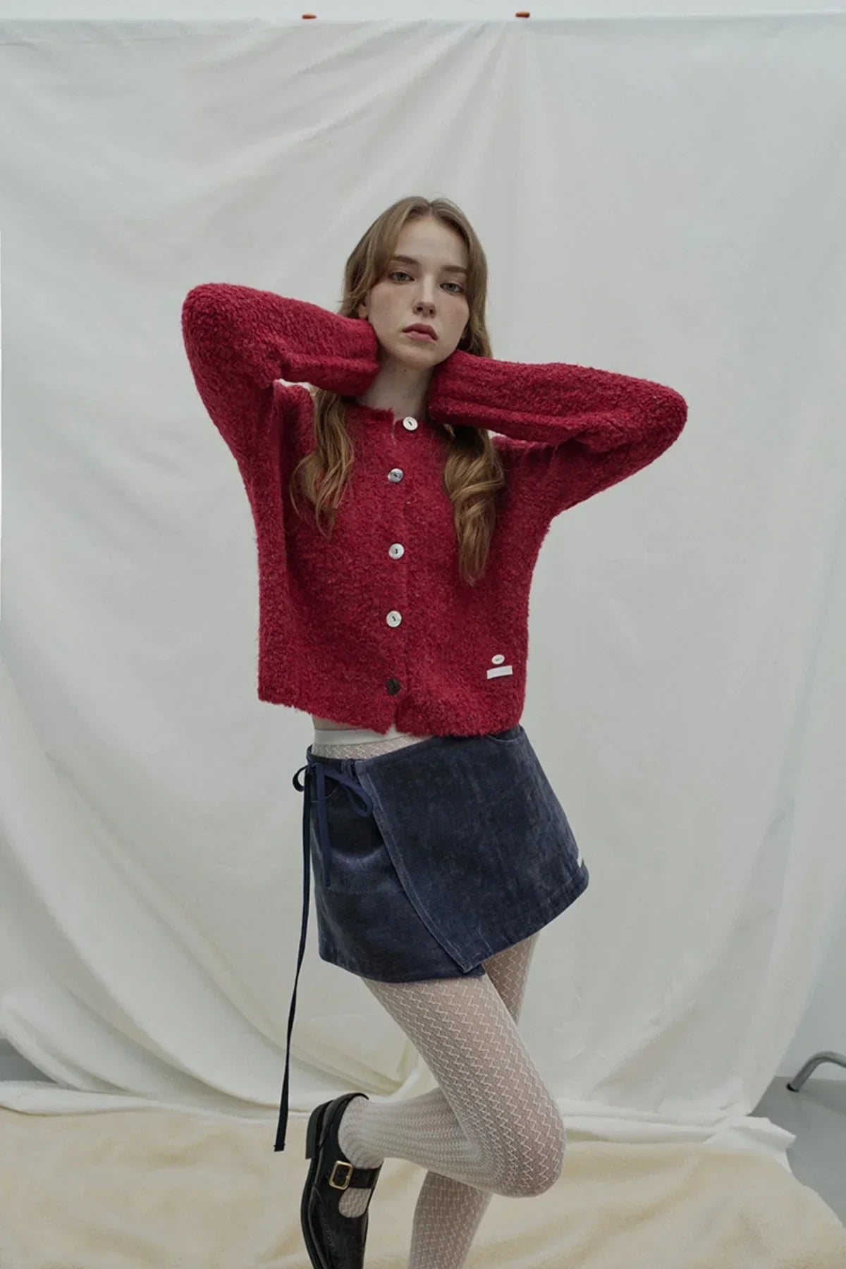 woman in red fluffy cardigan, navy blue velvet wrap skirt, patterned beige tights, and black shoes in studio