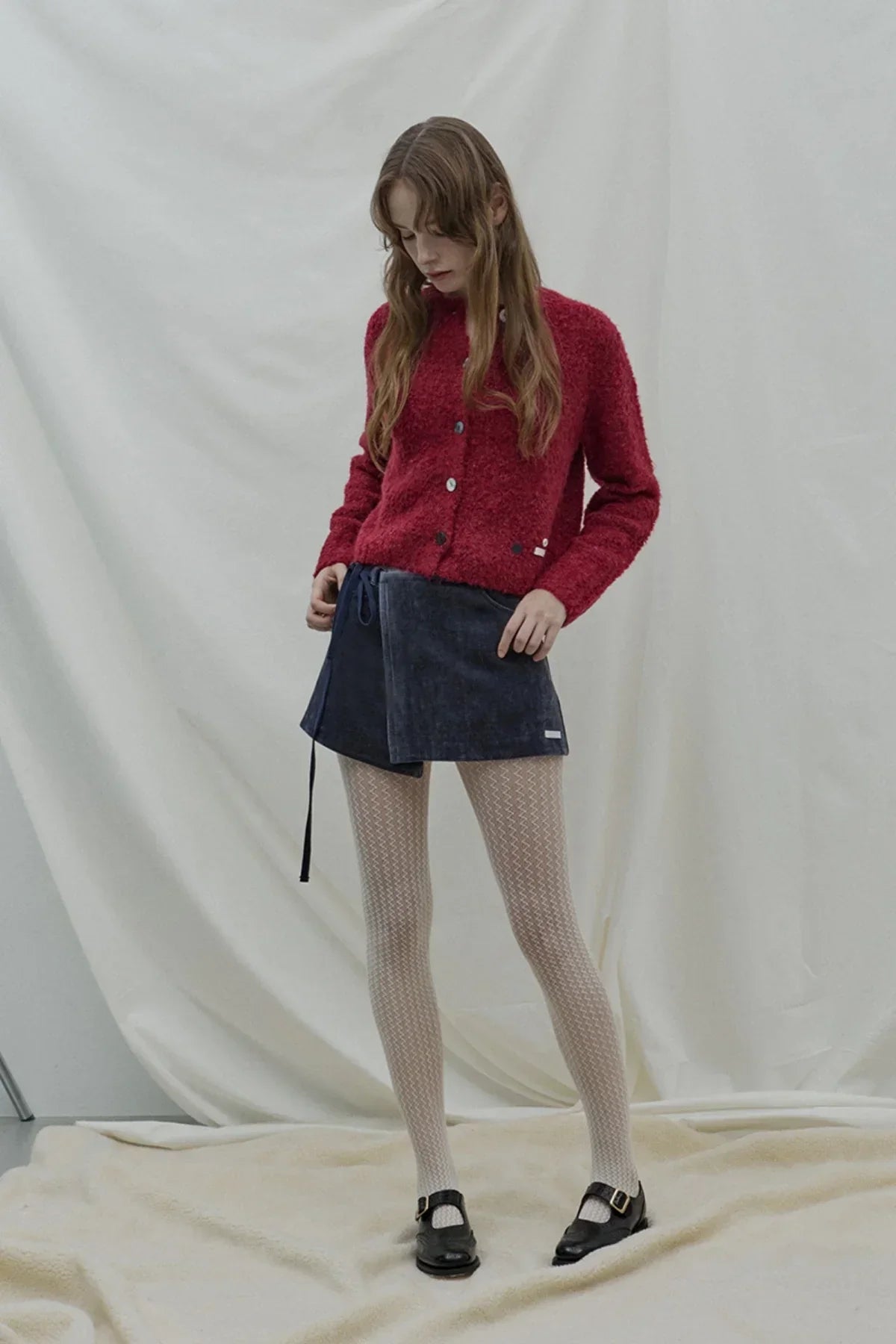 Woman wearing a red textured cardigan with a navy wrap skirt, patterned tights, and black buckle shoes