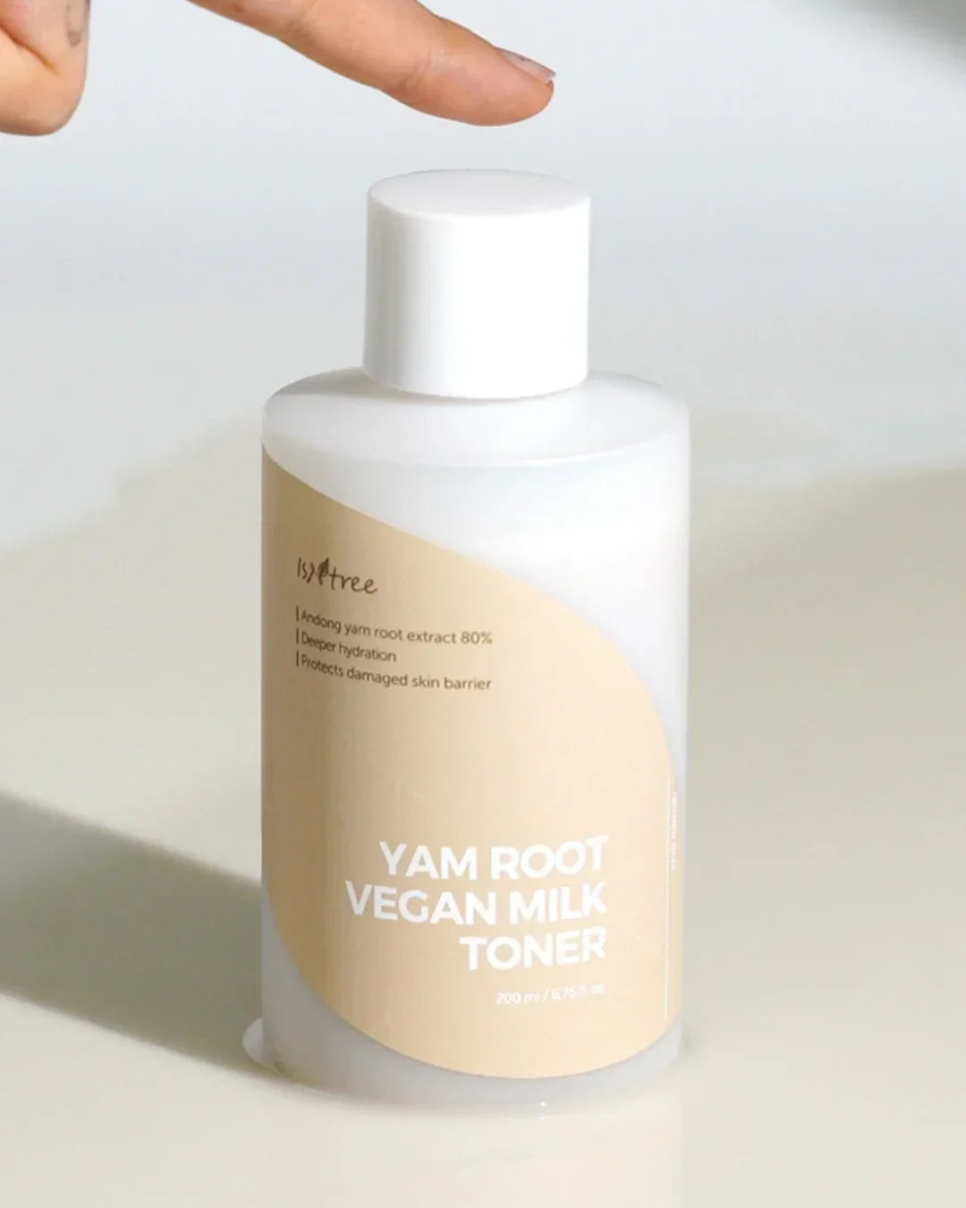 Bottle of Yam Root Vegan Milk Toner with beige label and white cap on light surface, finger pointing above