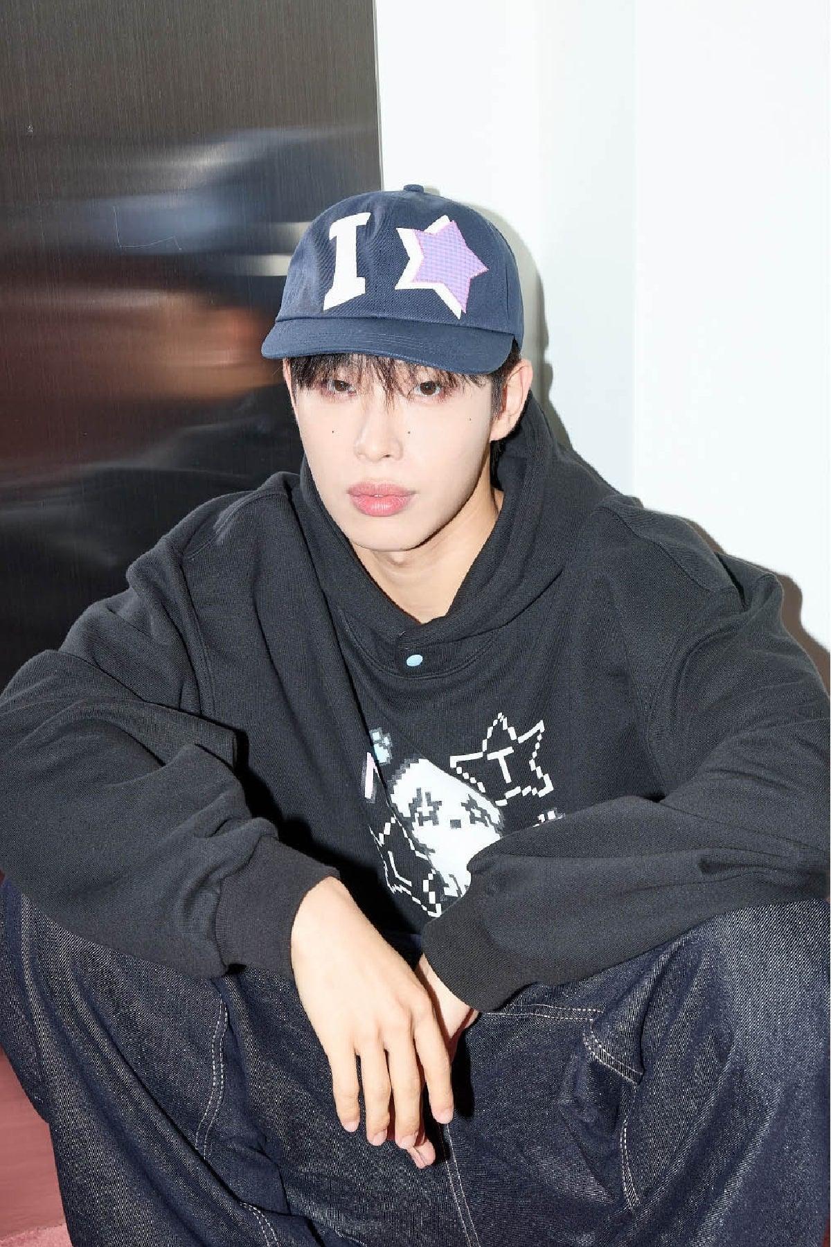 Young man in navy ball cap with star applique and black Korean streetwear hoodie, sitting casually.