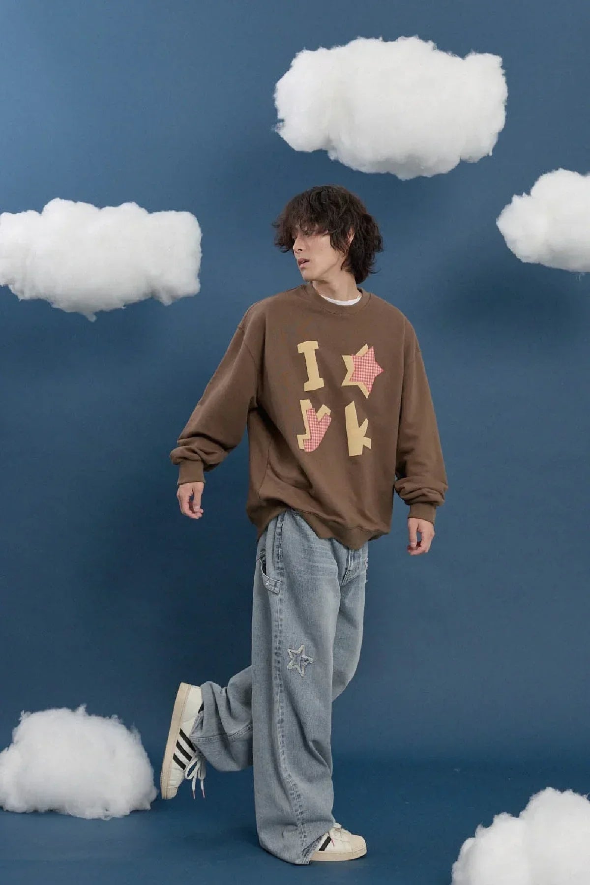 Model in brown YK check applique sweatshirt, loose jeans, and sneakers, posing with cloud props on a blue background, Korean fashion style from Malkn.