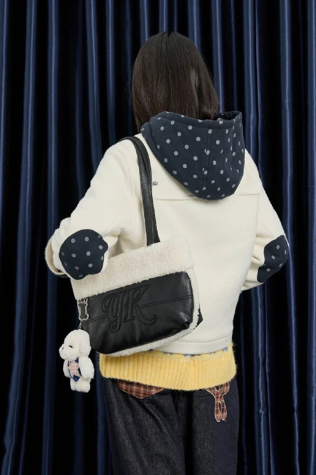 Woman with black and white shearling shoulder bag featuring YK logo and dog keychain, wearing polka dot hoodie and yellow sweater