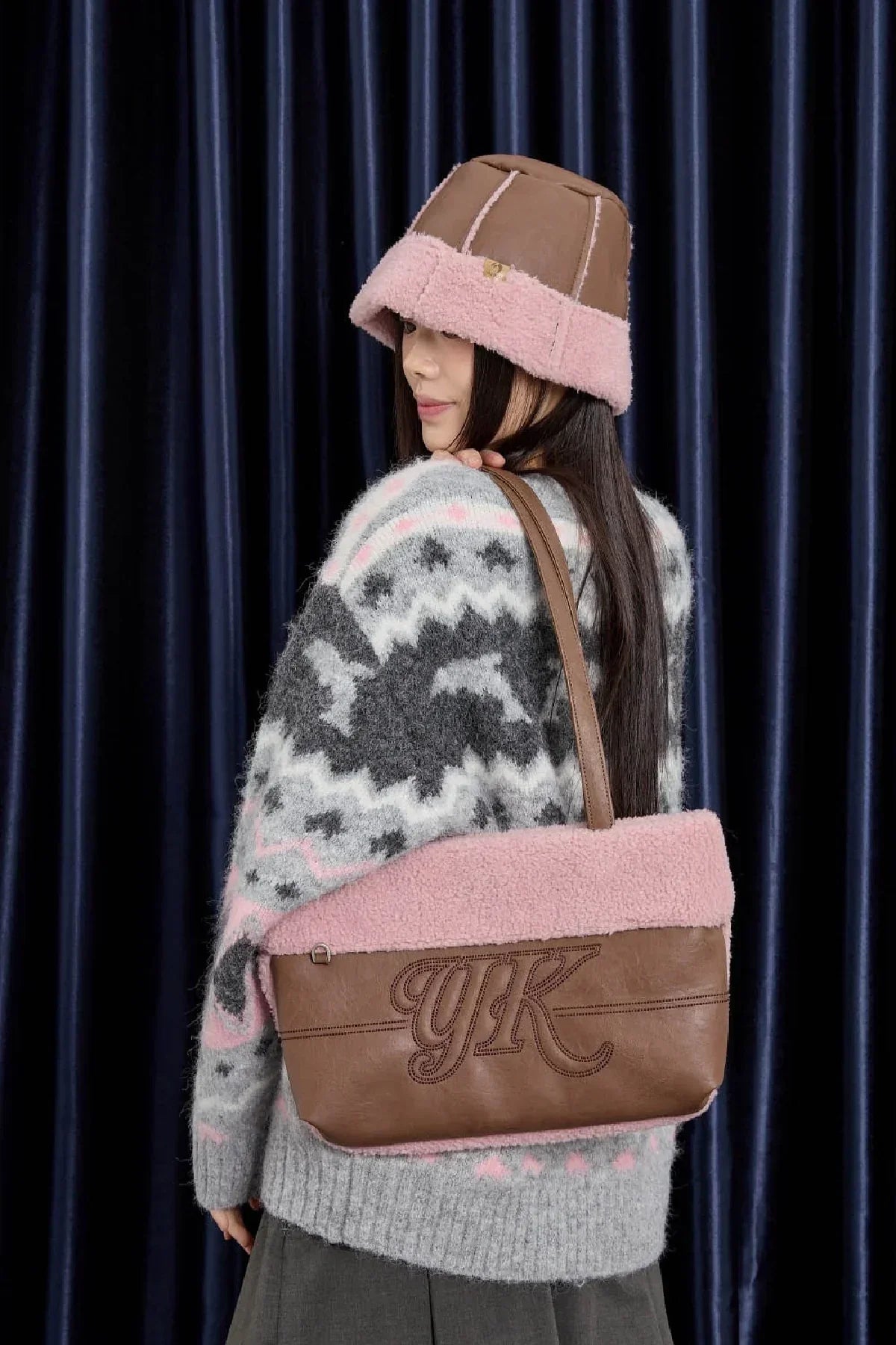 Woman wearing pink and brown bucket hat and gray patterned sweater, holding brown and pink shearling shoulder bag