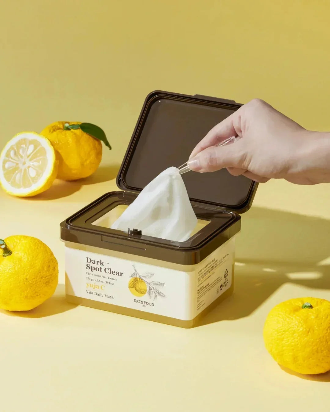 Hand pulling white sheet mask from dark spot clear Yuja C Vita Daily Mask box with yellow citrus fruits