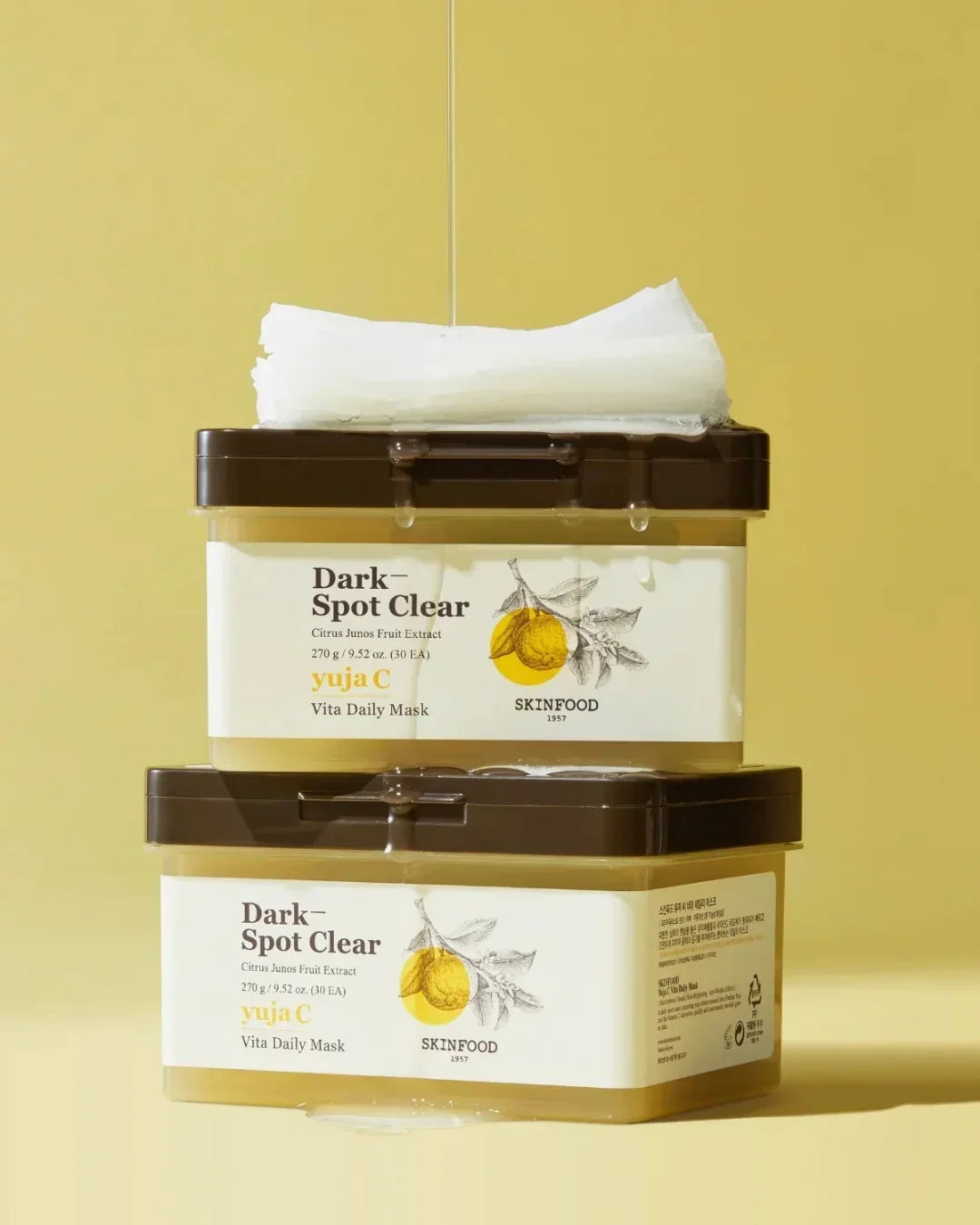 Two stacked Skinfood Yuja C Dark Spot Clear Vita Daily Mask containers with white mask sheets and pouring serum
