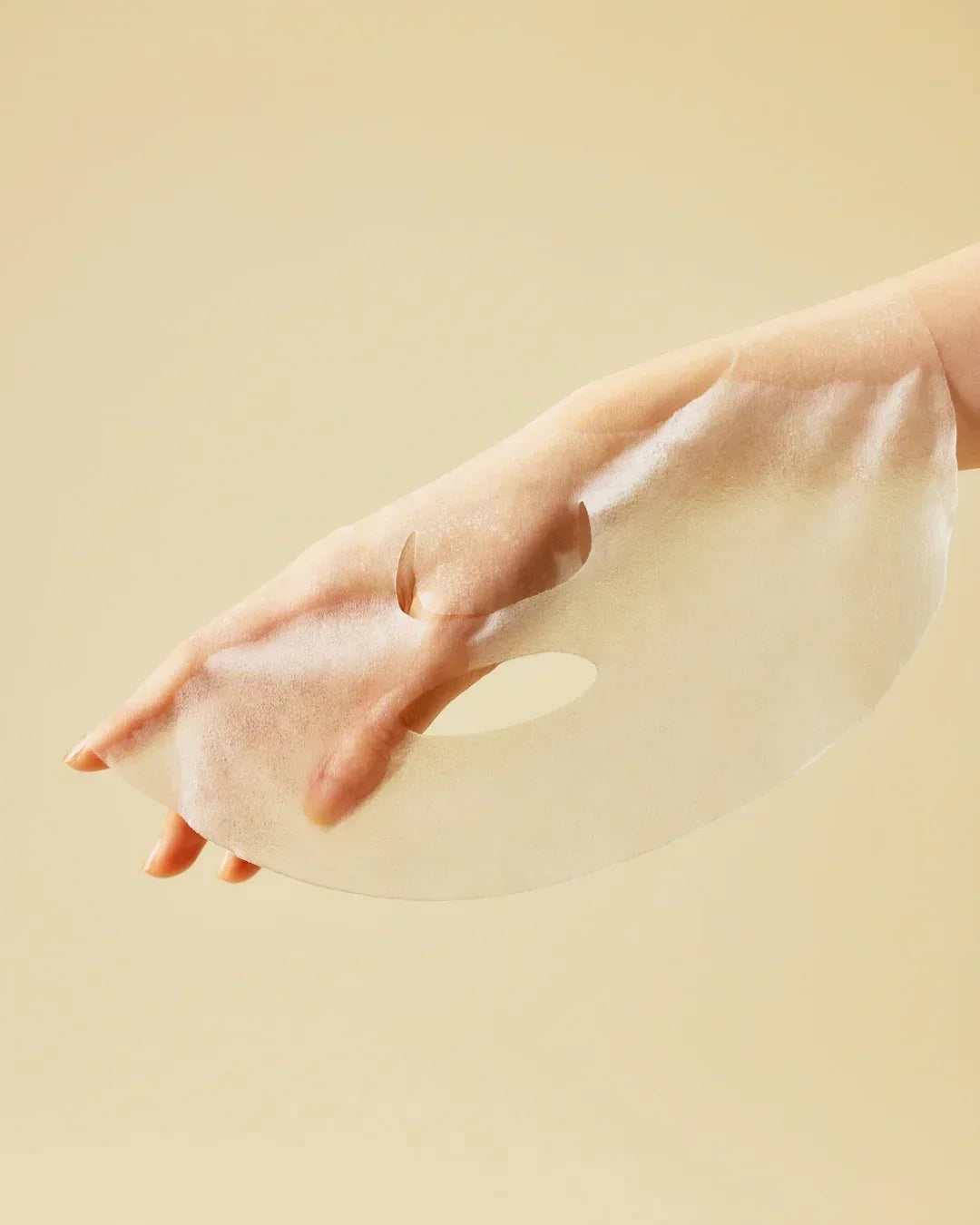 Hand holding translucent sheet face mask against beige background for skincare