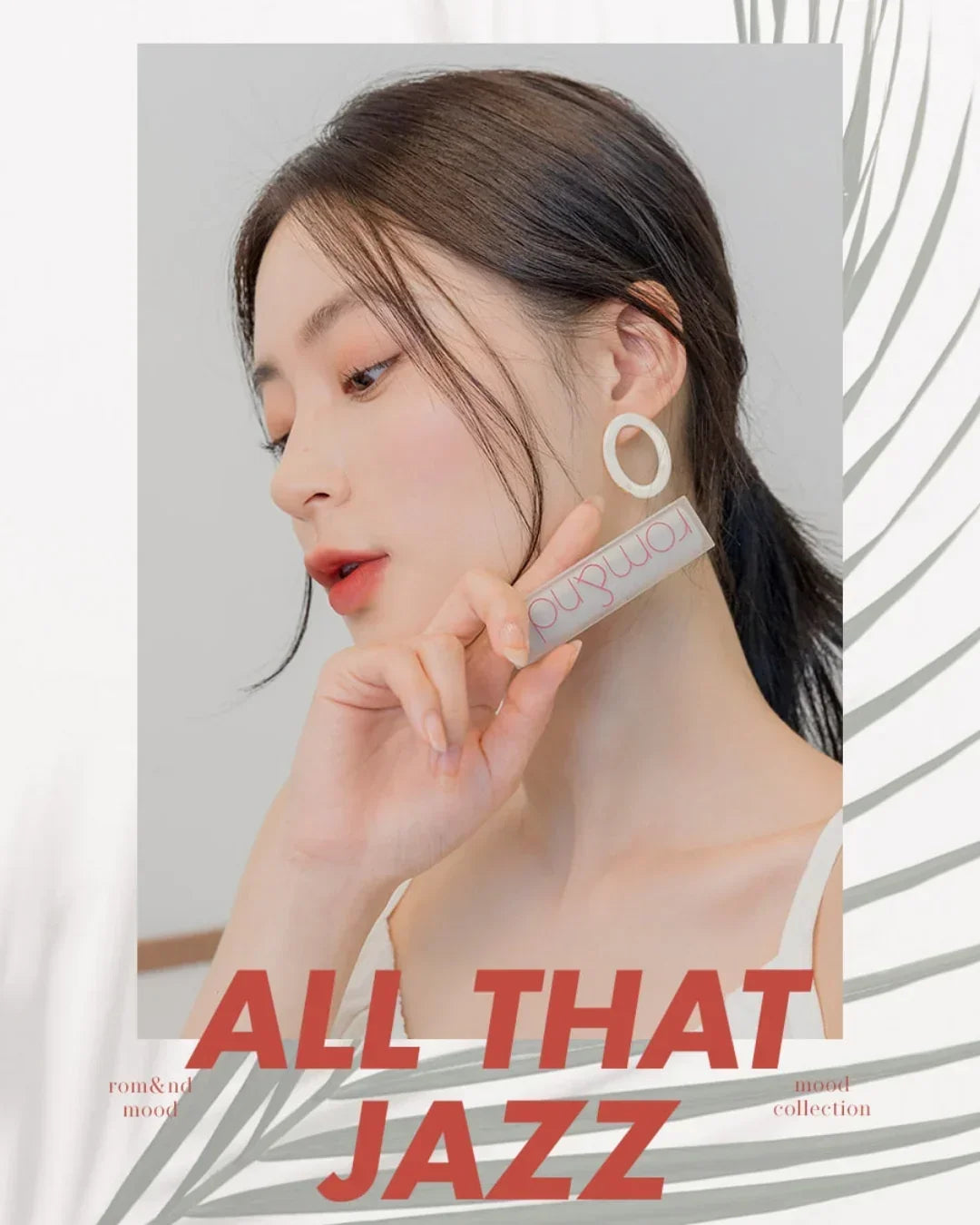 Close-up of woman with natural makeup holding Romand matte lipstick, wearing white tank top and circular earrings