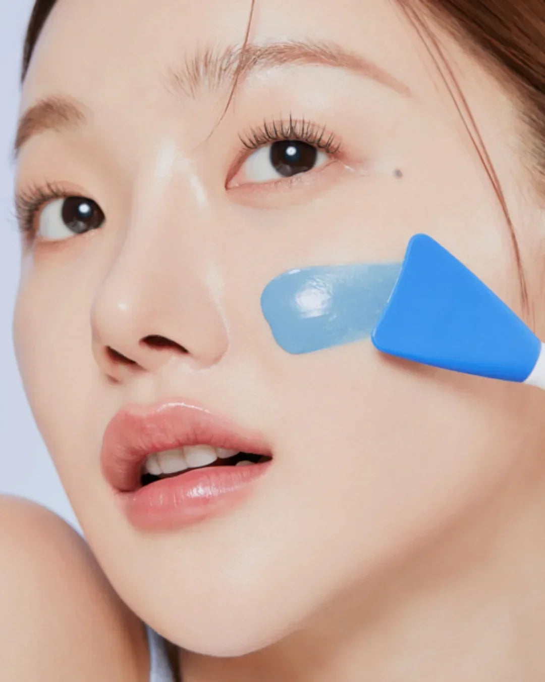 Close-up of woman applying blue face mask on cheek with silicone spatula for skincare