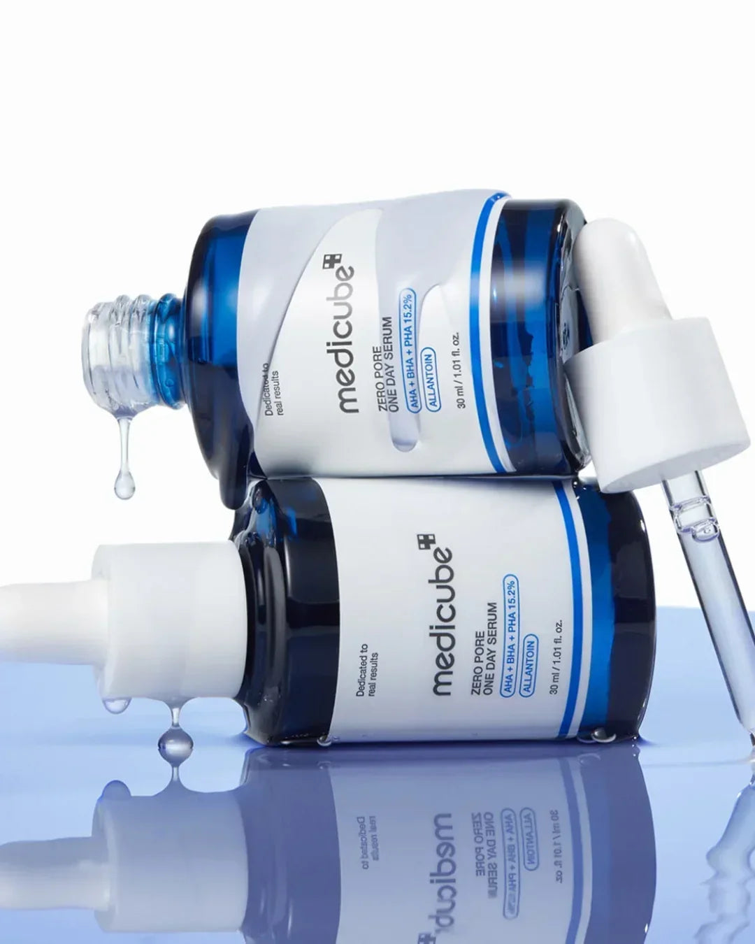 Two blue bottles of medicube Zero Pore One Day Serum with dropper caps and liquid drops on reflective surface