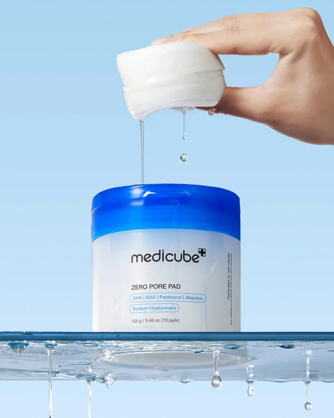 Hand holding wet medicube Zero Pore Pad with water dripping above blue and white container