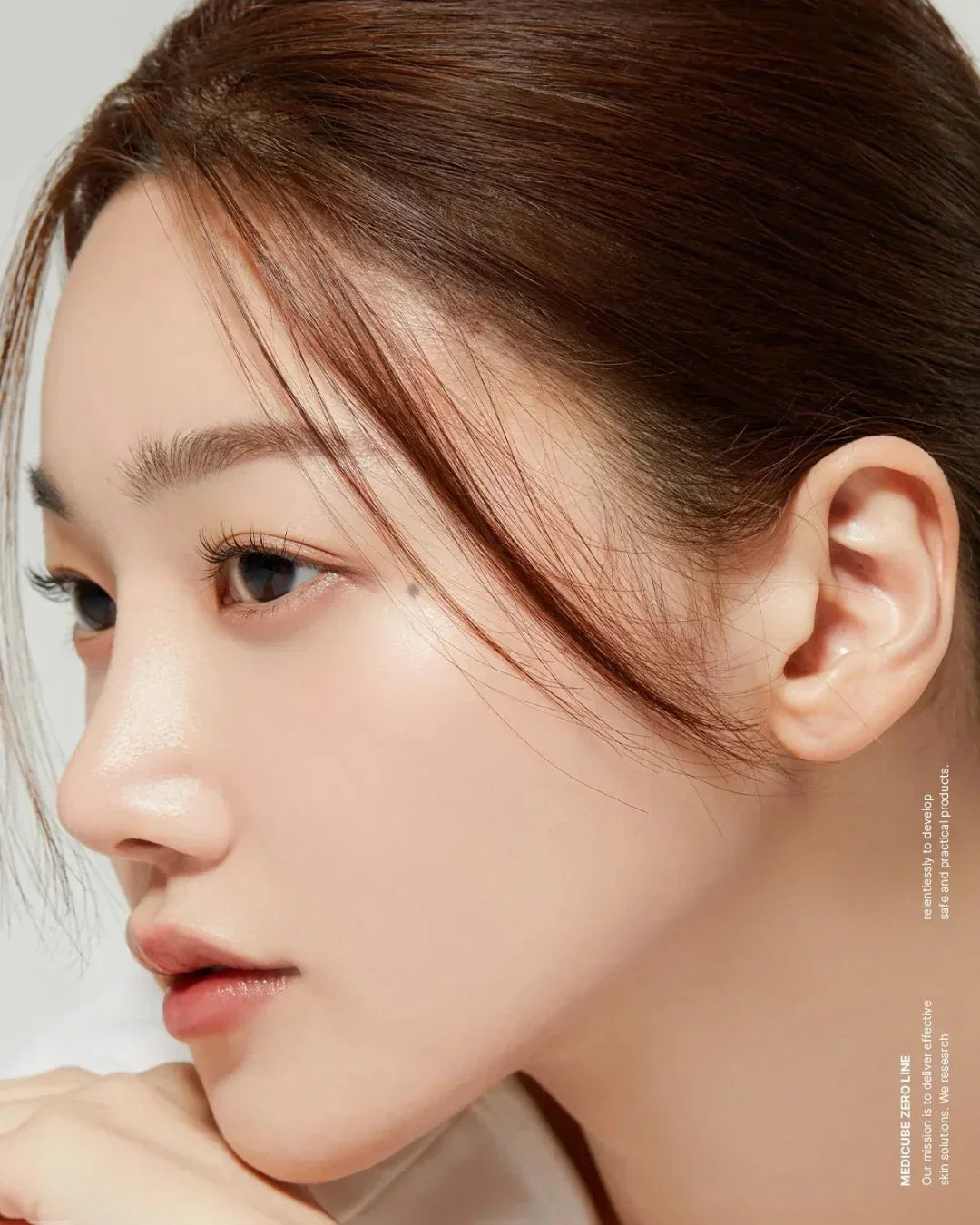Close-up profile of woman with clear skin, natural makeup, and brown hair against plain background