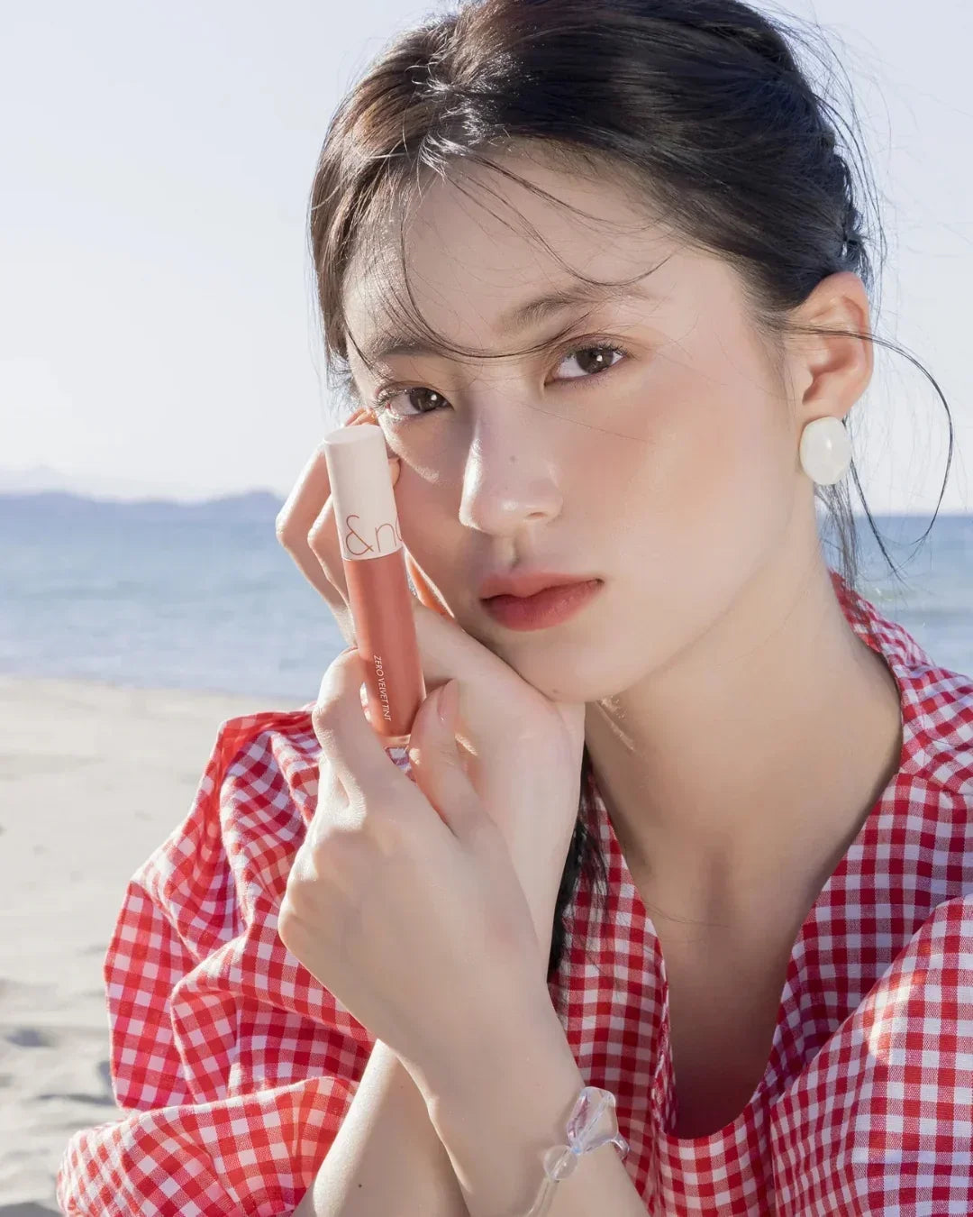 close-up of a woman in red checkered blouse holding Zero Velvet Tint lip product by the beach